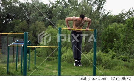 Hardy shirtless sportsman doing push ups on horizontal bar at nature. Strong and muscular guy training at sports ground. Young athletic guy exercising outdoor. Concept of active and healthy lifestyle 125939687