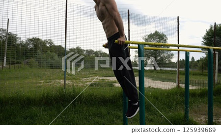 Young sportsman doing push ups on parallel bars at nature. Strong sporty man exercising at sports ground. Male shirtless athlete training hard outdoor. Concept of active sportive lifestyle. Slow mo Young sportsman doing push ups on parallel bars at nature. Strong sporty man exercising at sports ground. Male shirtless athlete training hard outdoor. Concept of active sportive lifestyle. Slow mo 125939689