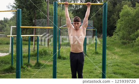 Muscular athlete doing pull ups on horizontal bar at sports ground. Young shirtless sportsman exercising at nature. Strong sporty man training outdoor. Concept of healthy and active lifestyle. Slow mo 125939720