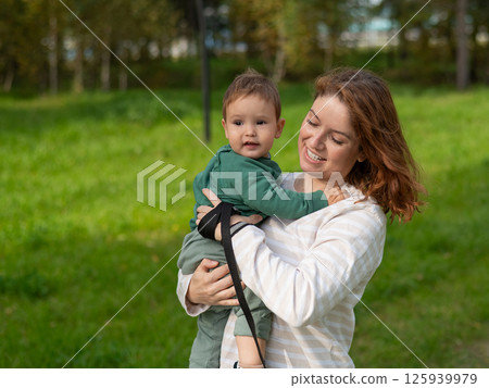 Caucasian woman holds her one-year-old son in her arms while walking in the park in autumn. Caucasian woman holds her one-year-old son in her arms while walking in the park in autumn. 125939979