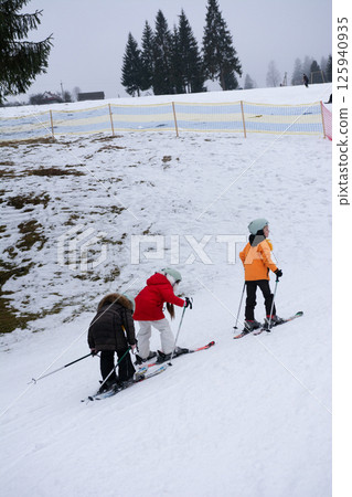 Children practice skiing on a snowy slope in a winter wonderland surrounded by pine trees under an overcast sky Children practice skiing on a snowy slope in a winter wonderland surrounded by pine trees under an overcast sky 125940935