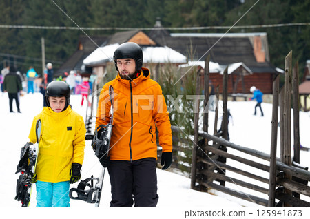 Snowy adventure with a father and child in vibrant winter attire exploring a snowy resort with snowshoes on a crisp, sunny day among pine trees 125941873