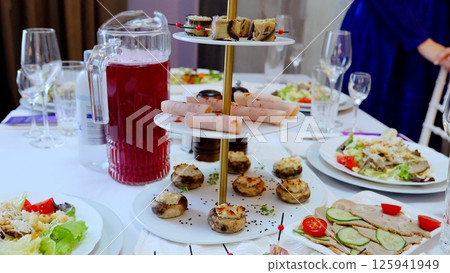 Festive table setting featuring diverse plates with salads, appetizers, cold cuts, stuffed mushrooms, accompanied by red juice pitcher and wine glasses 125941949