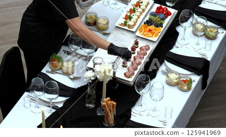 Waiter wearing black gloves arranging a variety of appetizers on an elegantly set table with a white tablecloth, black napkins, and candlelit decorations for a formal event Waiter wearing black gloves arranging a variety of appetizers on an elegantly set table with a white tablecloth, black napkins, and candlelit decorations for a formal event 125941969