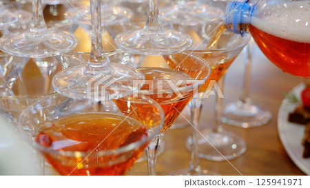 Bartender pouring vibrant orange cocktail into sleek glasses, creating sophisticated drink selection for upcoming social event Bartender pouring vibrant orange cocktail into sleek glasses, creating sophisticated drink selection for upcoming social event 125941971