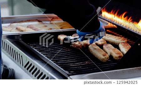 Professional chef wearing blue protective gloves, grilling sizzling sausages over flaming barbecue, carefully lifting meat from metal surface with professional tongs 125942025
