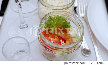 Gourmet caesar salad featuring succulent shrimp, cherry tomatoes, crisp lettuce, sprinkled parmesan in glass jar beside wine setting on white linen Gourmet caesar salad featuring succulent shrimp, cherry tomatoes, crisp lettuce, sprinkled parmesan in glass jar beside wine setting on white linen 125942066