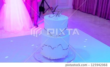 Elegant white wedding cake featuring silver monogram and pearl details, displayed under soft pink purple lighting with newlyweds subtly visible in background Elegant white wedding cake featuring silver monogram and pearl details, displayed under soft pink purple lighting with newlyweds subtly visible in background 125942068