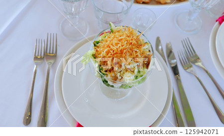 Green salad garnished with golden crispy onions, nestled in clear glass bowl, accompanied by elegant table setting with polished silverware and gleaming glassware Green salad garnished with golden crispy onions, nestled in clear glass bowl, accompanied by elegant table setting with polished silverware and gleaming glassware 125942071