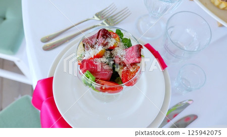 Hearty beef salad in glass bowl, layering crisp arugula, juicy tomatoes, creamy mozzarella, sharp parmesan over white plate with pink napkin, silverware nearby 125942075