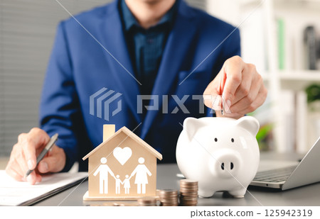 Family saving money concept. Businessman deposits coin into piggy bank with wooden house model, symbolizing family and love, family financial planning, and savings. cash management, life insurance, 125942319