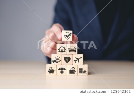 Insurance concept. Businessman arranges wooden blocks with insurance icons, financial security, health, home, and travel coverage. Top block features shield with checkmark, representing protection. 125942361