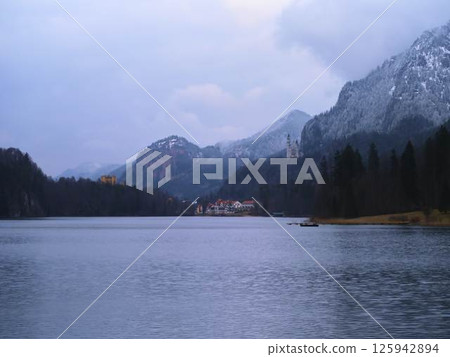 Snow-capped mountains overlook a serene lake with a castle, revealing a peaceful winter landscape at twilight 125942894