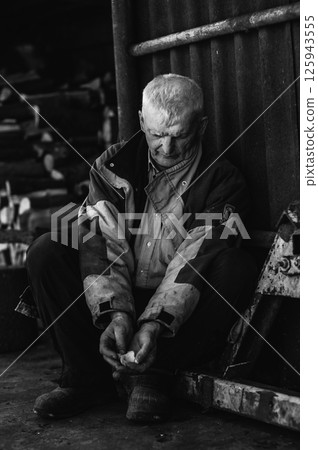 A pensioner rests on the ground, his gaze lost in thought. The elderly man wears clothes that have seen many years of work. A pensioner rests on the ground, his gaze lost in thought. The elderly man wears clothes that have seen many years of work. 125943555