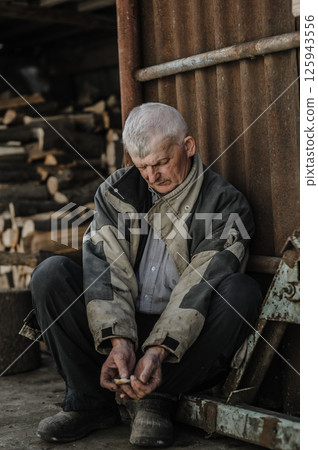 A pensioner rests on the ground, his gaze lost in thought. The elderly man wears clothes that have seen many years of work.  125943556