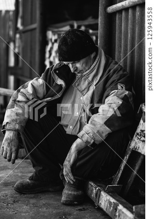 The pensioner crouches, exhausted by years of hardship. The elderly man is dressed in worn-out work clothes. The pensioner crouches, exhausted by years of hardship. The elderly man is dressed in worn-out work clothes. 125943558