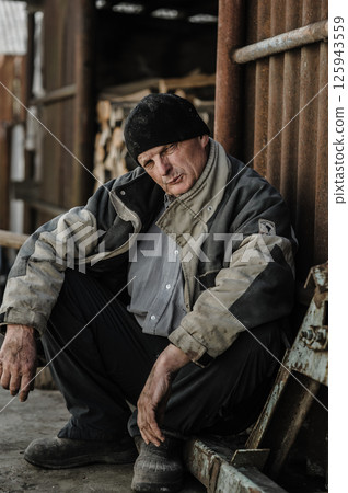 The pensioner crouches, exhausted by years of hardship. The elderly man is dressed in worn-out work clothes. 125943559