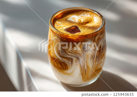 Iced coffee with milk swirls in glass on sunny white table 125943635
