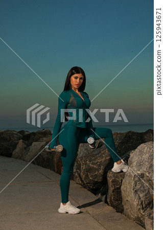 fitness enthusiast performs workout routine beachside promenade holding dumbbells and showcasing her strength against serene ocean backdrop. 125943671