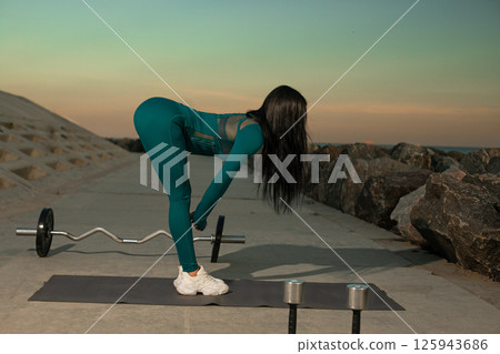 woman in fitted workout suit is engaged in deadlift exercise on yoga mat near beach. sunset illuminates horizon and creates serene atmosphere. 125943686