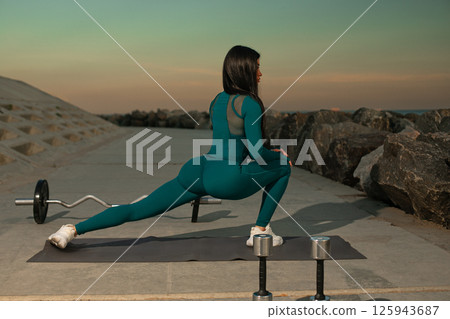 sporty woman performs stretching exercise on mat near rocky terrain as sun sets in background. She appears focused and ready for intense workout session after her warm up. 125943687