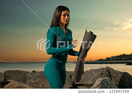 fitness enthusiast rolls out yoga mat on rocky shorelines as sun sets over horizon. tranquil setting offers peaceful ambiance perfect for meditation and stretching. 125943709