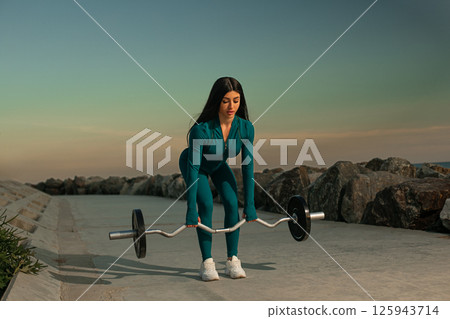 young woman in teal athletic outfit lifts barbell near rocky shore as sun sets. coastal workout captures serene yet empowering moment of fitness. young woman in teal athletic outfit lifts barbell near rocky shore as sun sets. coastal workout captures serene yet empowering moment of fitness. 125943714