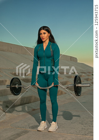 fitness enthusiast is lifting barbell in outdoor setting showcasing strength and focus. background highlights urban landscape warm hues of sunset illuminating scene. 125943715