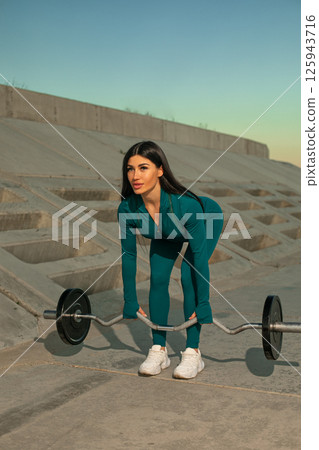 woman is engaged in deadlift exercise using barbell in urban setting sunrise. She is wearing fitness outfit and focused on her workout. surrounding area features concrete structures. 125943716