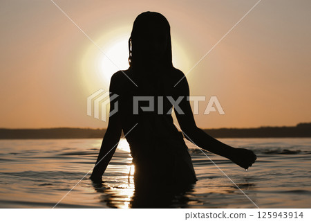 A silhouette of a teenager girl or young woman emerges from the water at sunset, capturing the serene beauty of nature and peaceful atmosphere. 125943914