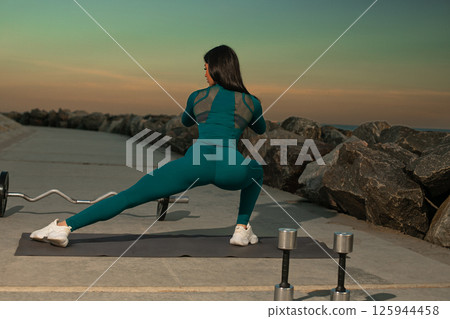 fitness enthusiast wearing teal outfit engages in lateral squat on mat placed on outdoor walkway lined rocks enjoying beautiful sunset. 125944458