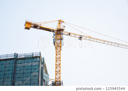 Yellow tower crane rising above skyscraper top under blue sky Yellow tower crane rising above skyscraper top under blue sky 125945544