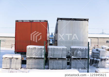 Two truck trailers in snowy warehouse yard with pallets and fence on clear winter day 125945621