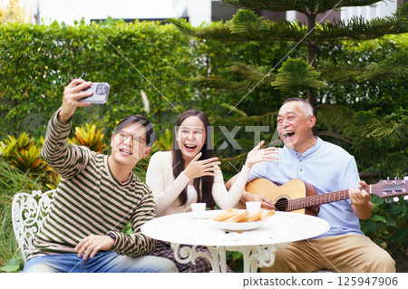 Happy Asian family spending time together, man and woman let their father playing a guitar and singing in backyard garden at home. Mental health care and wellbeing in senior adult. 125947906
