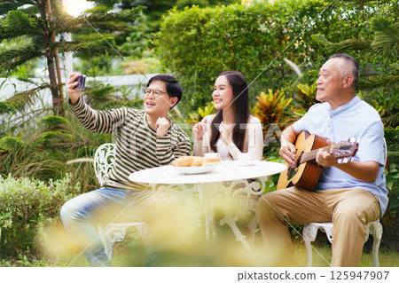 Happy Asian family spending time together, man and woman let their father playing a guitar and singing in backyard garden at home. Mental health care and wellbeing in senior adult. 125947907