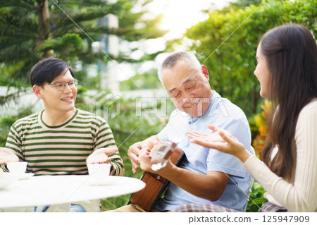 Happy Asian family spending time together, man and woman let their father playing a guitar and singing in backyard garden at home. Mental health care and wellbeing in senior adult. 125947909