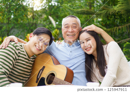Happy Asian family spending time together, man and woman let their father playing a guitar and singing in backyard garden at home. Mental health care and wellbeing in senior adult. Happy Asian family spending time together, man and woman let their father playing a guitar and singing in backyard garden at home. Mental health care and wellbeing in senior adult. 125947911