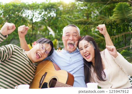 Happy Asian family spending time together, man and woman let their father playing a guitar and singing in backyard garden at home. Mental health care and wellbeing in senior adult. Happy Asian family spending time together, man and woman let their father playing a guitar and singing in backyard garden at home. Mental health care and wellbeing in senior adult. 125947912