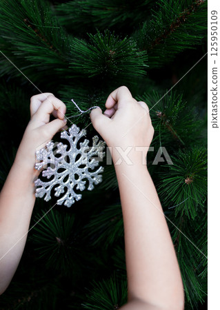 Child hanging silver snowflake ornament on green Christmas tree branches Child hanging silver snowflake ornament on green Christmas tree branches 125950109