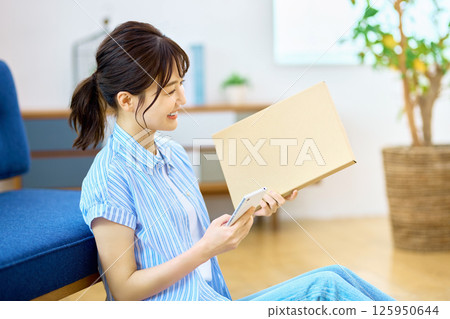 一位年輕女子拿著智能手機在客廳檢查郵件服務的行李 一位年輕女子拿著智能手機在客廳檢查郵件服務的行李 125950644