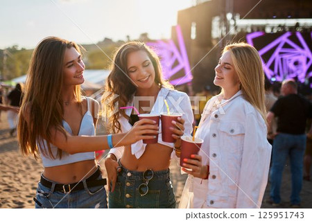 Women cheers with alcoholic beverages at sunset beach party. Smiling females holding cups, enjoying music event. Group of ladies celebrating, having fun on sandy shore near festival stage. Women cheers with alcoholic beverages at sunset beach party. Smiling females holding cups, enjoying music event. Group of ladies celebrating, having fun on sandy shore near festival stage. 125951743