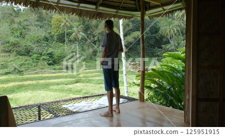 A young man enjoying scenic views while sipping coffee and standing on terrace of hut amidst the lush green landscape of a tropical jungle A young man enjoying scenic views while sipping coffee and standing on terrace of hut amidst the lush green landscape of a tropical jungle 125951915
