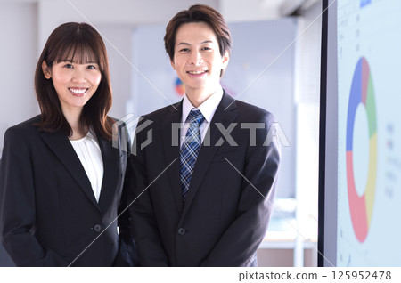 A man and a woman in suits standing in front of a screen in a conference room A man and a woman in suits standing in front of a screen in a conference room 125952478