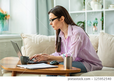 Focused woman in glasses working remotely on a laptop while sitting on a couch in a cozy home interior. Remote work and freelance lifestyle concept with modern home office setup. Focused woman in glasses working remotely on a laptop while sitting on a couch in a cozy home interior. Remote work and freelance lifestyle concept with modern home office setup. 125952540