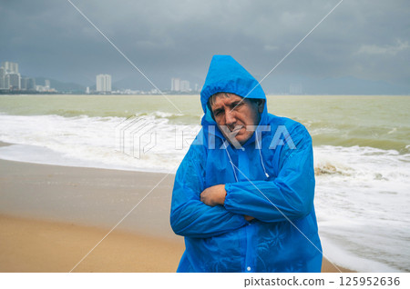 sad elderly man tourist in a raincoat on a beach by sea with waves in cloudy and rainy weather. Concept of the rainy and monsoon season 125952636