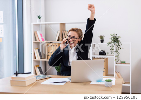 Middle-aged woman speaking on phone, celebrating with hand raised, working in modern office Middle-aged woman speaking on phone, celebrating with hand raised, working in modern office 125956193