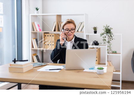 Confident adult woman wearing formal suit talking on phone and working on laptop, surrounded by office supplies. Bright office environment emphasizing productivity, multitasking, and work ethics. 125956194