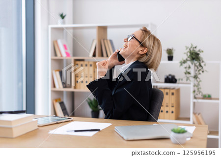 Woman enjoying phone conversation while seated at office desk in workplace demonstrating positivity. Middle-aged woman, professional attire, daytime setting with documentations, and greenery around 125956195