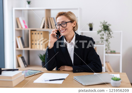 Professional woman in mid-thirties wearing business attire talking on phone at desk during work. 125956209
