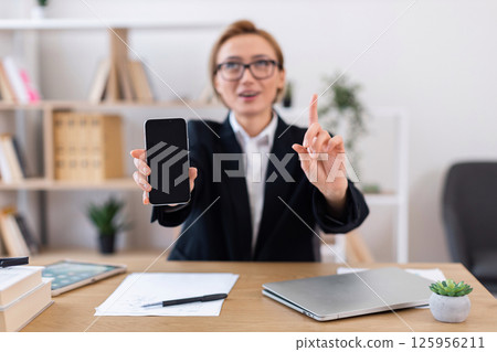 Businesswoman with glasses pointing while holding smartphone during professional discussion at office. Image portrays modern work, technology importance, communication in business environment. 125956211